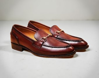 May include: A pair of brown leather loafers with gold-colored metal accents. The shoes have a classic design with a rounded toe and a low heel. The leather has a polished finish, and the shoes are set against a neutral background.