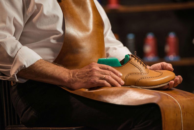 K&ouml;nnte beinhalten: Ein brauner Lederschuh im Brogue-Stil mit einem gr&uuml;nen Schuhspanner im Inneren, gehalten von einer Person, die eine braune Ledersch&uuml;rze und ein wei&szlig;es Hemd tr&auml;gt. Der Schuh hat dekorative Perforationen und braune Schn&uuml;rsenkel.