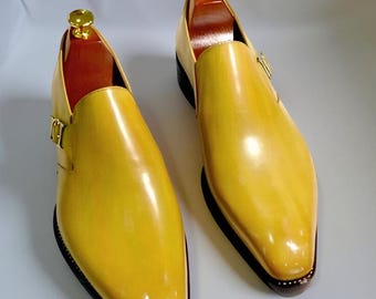 May include: A pair of yellow leather dress shoes with a shiny finish. The shoes have a pointed toe, a buckle closure, and a dark brown sole. Shoe trees are inserted inside the shoes. The shoes are displayed on a light gray surface.
