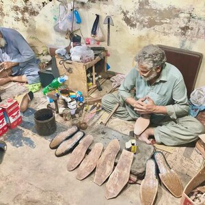 K&ouml;nnte beinhalten: Zwei Schuhmacher arbeiten in einer Werkstatt. Einer sitzt und bearbeitet eine Schuhsohle, w&auml;hrend der andere sitzt und an einem Lederst&uuml;ck arbeitet. Verschiedene Schuhkomponenten und Werkzeuge sind verstreut.