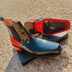 May include: Black and red Chelsea boots with a glossy finish. The boots have a square toe and a red sole. The heel and side panels are also red. The boots are displayed on a box.