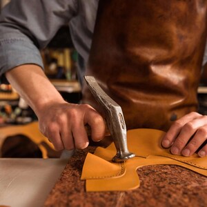 K&ouml;nnte beinhalten: Ein Handwerker bearbeitet gelbes Leder mit einem Hammer. Er tr&auml;gt eine braune Ledersch&uuml;rze und ein blaues Hemd. Die Werkstatt hat eine rustikale &Auml;sthetik. Das Leder wird auf einer braunen Oberfl&auml;che bearbeitet.