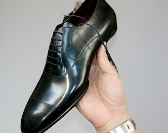 May include: A black leather dress shoe with a glossy finish. The shoe features a classic lace-up design and a rounded toe. The shoe is held up by a hand, showcasing its sleek and polished appearance. The shoe is a formal footwear item.