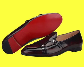 Può includere: Mocassini in pelle bordeaux con doppie fibbie argentate. La suola è rossa. Le scarpe sono esposte su sfondo giallo. Le scarpe hanno una finitura lucida.