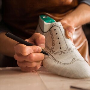 K&ouml;nnte beinhalten: Ein Schuhmacher zeichnet mit einem schwarzen Stift das Design auf einen wei&szlig;en Schuhleisten. Der Schuhleisten hat einen gr&uuml;nen Einsatz und ist mit dekorativen N&auml;hten und Perforationen versehen. Der Schuhmacher tr&auml;gt eine Ledersch&uuml;rze.