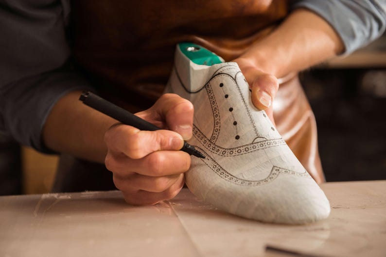 K&ouml;nnte beinhalten: Ein Schuhmacher zeichnet mit einem schwarzen Stift das Design auf einen wei&szlig;en Schuhleisten. Der Schuhleisten hat einen gr&uuml;nen Einsatz und ist mit dekorativen N&auml;hten und Perforationen versehen. Der Schuhmacher tr&auml;gt eine Ledersch&uuml;rze.