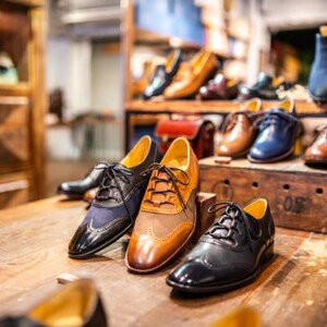 K&ouml;nnte beinhalten: Eine Auswahl an Herrenlederschuhen in verschiedenen Stilen und Farben. Die Abbildung zeigt Oxford-Schuhe, Stiefel und elegante Schuhe in Braun-, Schwarz- und Blaut&ouml;nen. Die Schuhe sind auf einer Holzoberfl&auml;che angeordnet.