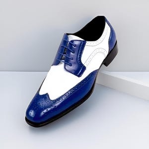 以下が含まれることがあります： 白と青のツートンカラーのオックスフォードシューズ。クラシックなウィングチップのデザインで、縁にブローグのディテールが施されています。靴紐とアッパーパネルは濃い青色で、白い部分と対照的です。ソールは黒色です。