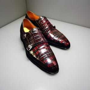 Puede incluir: Un par de zapatos de vestir de cuero burdeos con un patrón de piel de cocodrilo. Los zapatos tienen un cierre de doble hebilla con hebillas plateadas y una suela negra. El interior es de color naranja brillante.