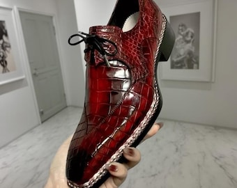 May include: A red leather shoe with a square toe and a textured, crocodile-like pattern. The shoe has a lace-up design and a small heel. The edges are trimmed with silver-colored material. The shoe is held by a person.
