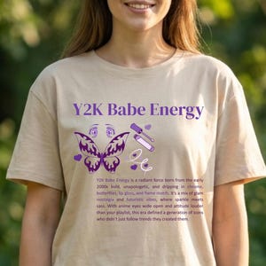 May include: Beige t-shirt featuring the text "Y2K Babe Energy" in purple, accompanied by butterfly and makeup illustrations. The shirt has a round neck and short sleeves. The text below describes the Y2K aesthetic.