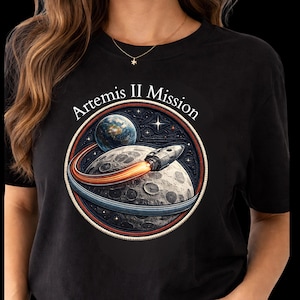 Puede incluir: Camiseta negra con un diseño circular que dice "Artemis II Mission". El diseño muestra un cohete despegando de una luna, con la Tierra y estrellas en el fondo. El diseño utiliza elementos rojos, naranjas, azules y blancos.