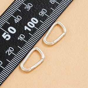 May include: Two silver-colored, D-shaped carabiner clips next to a black ruler with white markings. The ruler shows measurements in inches and centimeters. The background is a solid tan color.