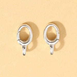 Puede incluir: Dos cierres ovalados plateados con un pequeño gancho en la parte inferior, sobre un fondo amarillo claro. Estos cierres son probablemente para joyas o pequeños accesorios. Diseño sencillo y funcional.