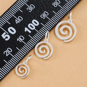Puede incluir: Tres colgantes plateados en espiral de diferentes tamaños sobre un fondo beige. Una regla negra con marcas blancas se coloca en diagonal, sirviendo de escala. Los colgantes presentan un diseño en espiral y un pequeño lazo para sujetar. Medidas en centímetros.