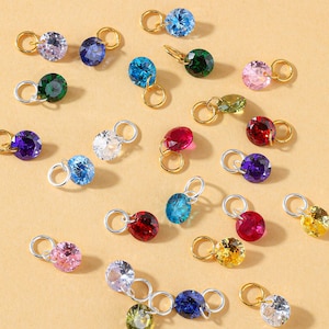 May include: A selection of gemstone charms in various colours, with gold and silver-tone findings. The charms feature blue, green, pink, red, purple, and yellow gemstones. Each charm has a round, faceted gemstone.