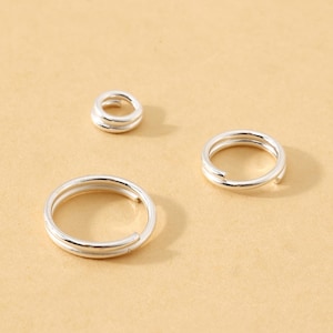 May include: Three silver-colored split rings of varying sizes are arranged on a tan surface. The rings are made of a shiny metal and have a simple, circular design. The smallest ring is a stack of two rings, while the other two are single rings.