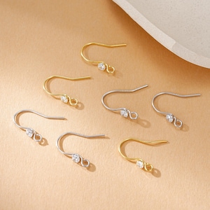 Può includere: Una collezione di ganci per orecchini color oro e argento con piccole gemme trasparenti. I ganci sono disposti su una superficie marrone chiaro, mostrando il loro design delicato e le varie finiture metalliche. Le gemme aggiungono un tocco di brillantezza ai componenti dei gioielli.