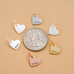 May include: Heart-shaped charms in silver, gold, and rose gold, each with a loop and the name "Mikre". The charms are shown with a coin for scale. The charms are suitable for jewellery making.
