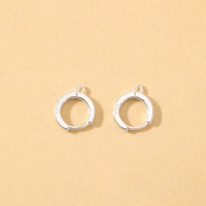 以下が含まれることがあります： 小さなループが付いた2つのシルバーフープイヤリング。イヤリングは丸く、ヒンジ留めです。金属は光沢のある仕上げです。明るいタン色の背景に表示されています。
