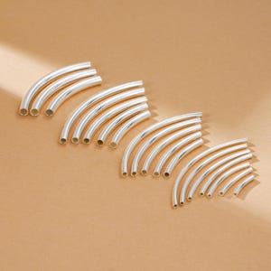 May include: Several sets of silver-colored, curved metal tubes are arranged on a light brown surface. The tubes are hollow and have a smooth, reflective finish. The varying sizes suggest they are components for jewelry or crafts.