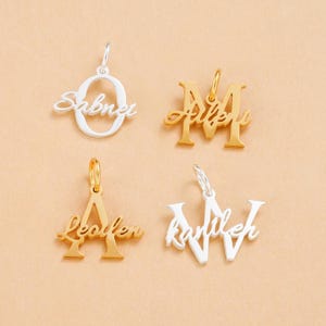 Könnte beinhalten: Vier personalisierte Buchstabenanhänger in Gold und Silber. Die Anhänger haben die Form der Buchstaben O, M, A und W, mit den Namen Sabrina, Allie, Lealfer und Kaylileh in Schreibschrift. Jeder Anhänger hat eine kleine Schlaufe zum Befestigen an einer Kette.