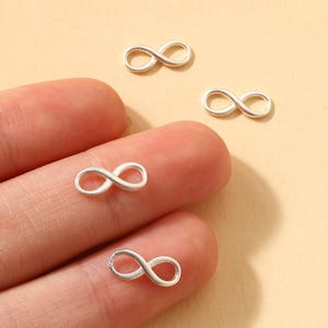 Könnte beinhalten: Nahaufnahme von mehreren kleinen, silberfarbenen Unendlichkeitszeichen-Anhängern. Die Anhänger sind auf einer hellbraunen Oberfläche verstreut, einige liegen auf den Fingerspitzen einer Person. Die Anhänger sind aus glänzendem Metall.