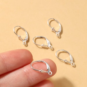 May include: Several silver-colored leverback earring findings are displayed against a light tan background. One is held by a person's fingers. The findings have a hinged design with a small loop for attaching earrings. These are jewelry making supplies.