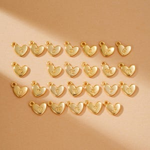 May include: A collection of gold-colored heart-shaped charms, each with a different letter of the alphabet engraved on its surface. The charms are arranged in rows and columns against a neutral background. Each charm has a small loop at the top for attaching to a chain or bracelet.