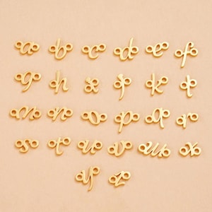 May include: Gold-coloured alphabet charms, each with a loop on either side for attaching to jewellery. The letters are in a cursive style, with a variety of fonts. The charms are arranged on a light tan surface.