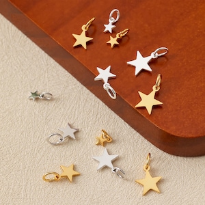 Puede incluir: Colección de colgantes en forma de estrella en tonos dorados y plateados. Los colgantes son pequeños, con un diseño de estrella de cinco puntas, y están unidos a pequeños aros. Los colgantes se exhiben sobre una superficie de madera marrón.