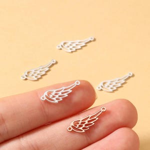 May include: Several small, silver-colored wing charms with intricate feather detailing. Each charm has a small loop at the top for attaching to jewelry. The charms are displayed against a light peach background.