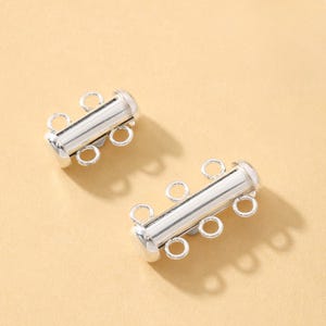 May include: Two silver-colored jewelry clasps with three loops on each side. The clasps have a cylindrical center with a rounded end. The background is a light tan color.