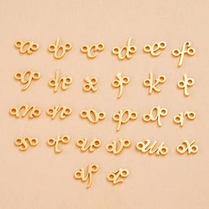May include: A collection of gold-coloured metal charms shaped like lowercase letters of the alphabet. Each letter features a cursive, stylised design with small circular loops at the ends of the strokes, arranged in rows on a plain background.