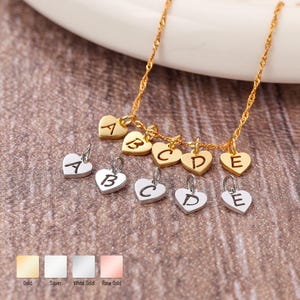 May include: A gold-coloured twisted chain necklace featuring five dangling heart-shaped charms engraved with the letters A, B, C, D, and E. Below the necklace are five silver-coloured heart-shaped charms also engraved with the letters A, B, C, D, and E.
