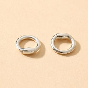 以下が含まれることがあります： 2つの銀色の、不規則な形のリング。リングは滑らかで磨かれた仕上げで、わずかに不均一な表面を持っています。デザインはシンプルでモダンで、ジュエリーやクラフトプロジェクトに適しています。リングは明るいベージュの背景に表示されています。