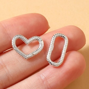 Könnte beinhalten: Zwei silberfarbene Schmuckverschlüsse. Einer ist herzförmig, der andere ein rechteckiges Oval. Beide Verschlüsse haben ein strukturiertes, gedrehtes Seildesign. Die Verschlüsse werden in einer Hand vor einem hellbraunen Hintergrund gehalten.