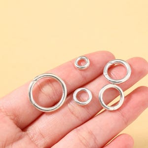 May include: A collection of silver-colored, circular clasps of varying sizes. The clasps are open, revealing a small hinge mechanism. The clasps are displayed against a light yellow background.