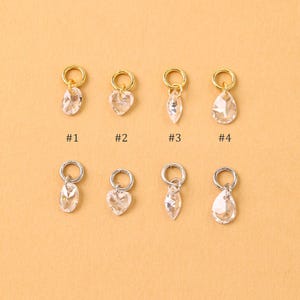 Könnte beinhalten: Acht Kristallanhänger mit goldenen und silbernen Ringen werden gezeigt. Die Anhänger haben vier Formen: oval, Herz, Blatt und Tropfen. Jeder Anhänger ist mit einer Nummer von #1 bis #4 versehen. Der Hintergrund ist einfarbig hellbraun.