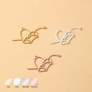 Könnte beinhalten: Drei herzförmige Metallanhänger in Gold, Silber und Roségold. Jeder Anhänger hat ein Herzdesign mit einem Schlaufenmuster und einer kleinen Schlaufe am Ende zum Befestigen. Die Anhänger werden auf einem hellbeigen Hintergrund präsentiert.