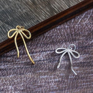 Peut inclure: Deux pendentifs en forme de nœud papillon, l'un doré et l'autre argenté, sont présentés sur une surface sombre texturée. Le pendentif doré a une boucle en haut et le pendentif argenté a une boucle en haut. Ces pendentifs sont probablement pour la fabrication de bijoux.