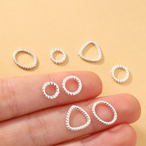 May include: A collection of small, silver-colored jewelry findings in various shapes, including circles, ovals, and triangles. The findings have a twisted rope-like texture and are displayed against a light tan background.