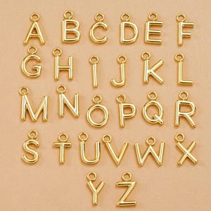 Könnte beinhalten: Eine Sammlung goldfarbener Alphabet-Anhänger, jeweils mit einer Schlaufe oben zum Befestigen an Schmuck oder anderen Bastelarbeiten. Die Buchstaben A bis Z werden angezeigt und zeigen eine Vielzahl von Schriftarten und Stilen.