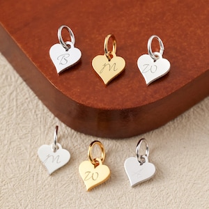 May include: A selection of heart-shaped charms in silver and gold. Each charm has a small loop for attachment and features a single initial or number. The charms are displayed on a wooden surface.