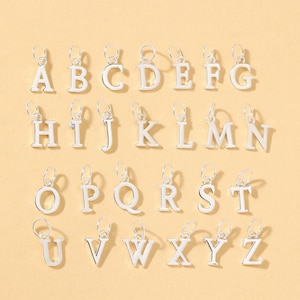 May include: Silver alphabet charms, each letter from A to Z, are displayed against a light yellow background. The charms are in a classic serif font, with a small loop at the top for attaching to a chain or bracelet.