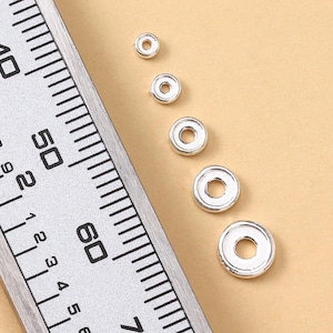 May include: Close-up of several small, silver-colored, circular beads with a hole in the center. A metal ruler with black markings and numbers is in the frame, providing scale. The beads are arranged on a light brown surface.