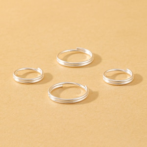 May include: Four silver-toned rings, each featuring a double-band design with a small gap at the top. The rings have a smooth, polished finish and a circular shape, arranged in a scattered formation on a solid, light tan-coloured surface.