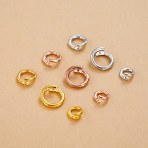 May include: An assortment of jump rings in gold, silver, and rose gold hues. The open rings are displayed on a neutral backdrop, highlighting their varied sizes and finishes. These jewellery findings are used for connecting jewellery components.