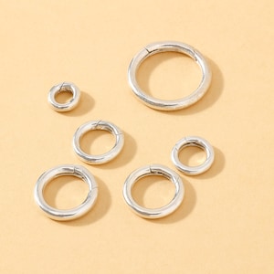 May include: A collection of silver-colored metal clasps in various sizes. The clasps are circular with a hinged opening, designed for jewelry making or crafting. The clasps are displayed on a light tan surface.