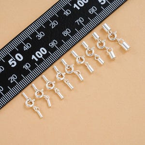 Op de afbeelding: Een verzameling zilverkleurige sieraden sluitingen met veersluitingen en cilindrische uiteinden. Op de achtergrond een zwarte liniaal met witte markeringen. De sluitingen zijn gerangschikt op een lichtbruin oppervlak.